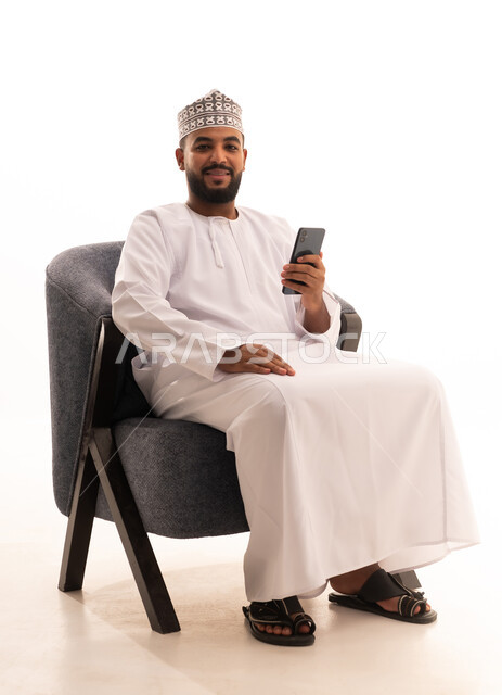 إجراء مكالمة فيديو عبر الجوال، التواصل مع الأهل والاصدقاء، تصفح مواقع التواصل الاجتماعي، استخدام جهاز تقني حديث،بورتريه لرجل عربي خليجي عماني يرتدي الدشداشة والكمة يجلس على كرسي ويعمل على الهاتف المحمول، خلفية بيضاء
