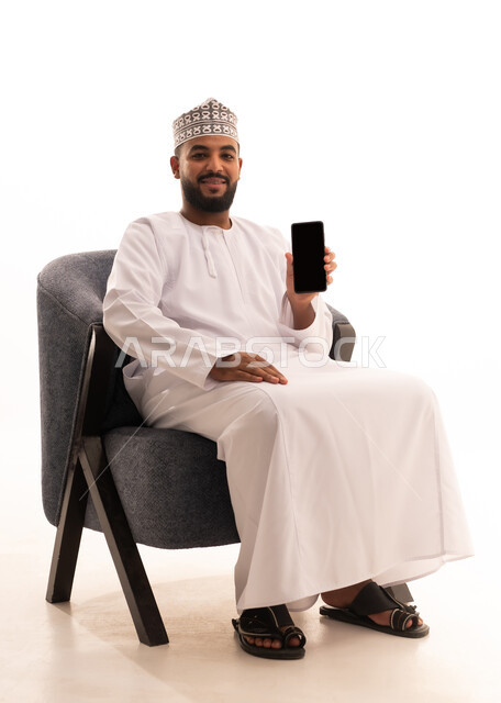 ايماءات السعادة و السرور، مواكبة التقدم العصري الحديث، مفهوم التطورات التقنية والتكنولوجية، بورتريه لرجل عربي خليجي عماني يرتدي الدشداشة والكمة يجلس على كرسي يقوم بعرض شاشة سوداء فارغة عبر الهاتف المحمول ، خلفية بيضاء