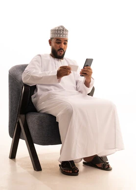 مفهوم الدفع والتسديد عن بعد عبر تطبيقات الجوال، ايماءات المفجأة و الذهول، استخدام البطاقة البنكية الالكترونية ، تطور التكنولوجيا والأجهزة التقنية،بورتريه لرجل عربي خليجي عماني يرتدي الدشداشة والكمة يجلس على كرسي يستخدام الهاتف المحمول و يمسك بطاقة بيضاء، خلفية بيضاء
