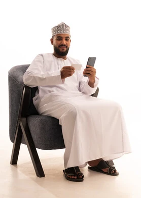 The use of electronic bank cards, the concept of remote payment and settlement through mobile applications, the evolution of technology and technical devices, a portrait of an Arab Gulf Omani man wearing a dishdasha and kumma sitting on a chair using a mobile phone and holding a white card, white background.