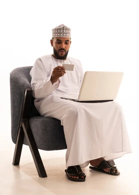 The use of electronic bank cards, gestures of surprise and astonishment, the concept of remote payment and settlement through laptop applications, the evolution of technology and technical devices, a portrait of an Arab Gulf Omani man wearing a dishdasha and kumma sitting on a chair working on a laptop and holding a white card, white background.