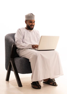 Remote business management, using modern and advanced devices, a portrait of an Arab Gulf Omani man wearing a dishdasha and kumma sitting on a chair using a laptop, integrating technology with work, completing required tasks via the laptop, white background.