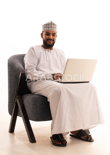 The use of modern and advanced devices, remote business management, a portrait of an Arab Gulf Omani man wearing a dishdasha and kumma sitting on a chair using a laptop, integrating technology with work, completing required tasks via the laptop, white background.