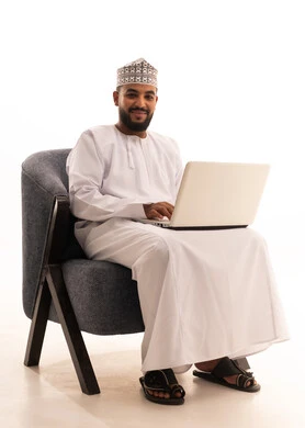 The use of modern and advanced devices, remote business management, a portrait of an Arab Gulf Omani man wearing a dishdasha and kumma sitting on a chair using a laptop, integrating technology with work, completing required tasks via the laptop, white background.