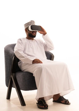 Spending leisure time in an enjoyable way, seeing imaginary scenes, interacting and immersing oneself in virtual reality, using modern technological devices, a portrait of an Arab Gulf Omani man wearing a dishdasha and kumma sitting on a chair wearing VR glasses, white background.