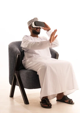 التطور التقني والتكنولوجي، الاندماج ومحاكاة العالم الخيالي، المتعة والتسلية بالمشاهدة عبر نظارة الواقع الافتراضي، بورتريه لرجل عربي خليجي عماني يرتدي الدشداشة والكمة يجلس على كرسي يضع نظارات ثلاثية الأبعاد (الفي ار)، خلفية بيضاء