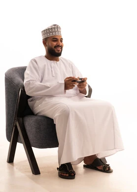 Enjoying modern technologies and technology, the concept of fun and entertainment, gestures of immersion and interaction, a portrait of an Arab Gulf Omani man wearing a dishdasha and kumma sitting on a chair holding a game controller, having a good time playing video games, white background.