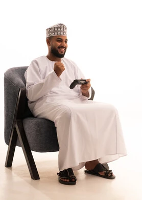 رفع قبضة اليد للاعلى بتعابير الفوز و الربح, اجهزة وتقنيات الكترونية حديثة ترفيهيه, قضاء وقت ممتع ومسلي، التفاعل والاندماج مع ألعاب الفيديو والبلايستيشن،  بورتريه لرجل عربي عماني خليجي يرتدي دشداشة وكمة يجلس على كرسي ويحمل بيده عصا التحكم، خلفية بيضاء