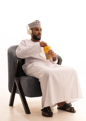 انشطة ترفيهية منزلية، ايماءات وتعابير التعجب والصدمة، الاستعداد لمشاهدة الافلام وإحضار السناكات اللذيذة، مشاهدة فلم سينمائي، بورتريه لرجل عربي عماني خليجي يرتدي الدشداشة و الكمة و سماعات الهيدفون يجلس على كرسي يحمل بيده علبة الفوشار، خلفية بيضاء