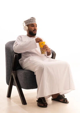 ايماءات وتعابير التعجب والصدمة، الاستعداد لمشاهدة الافلام وإحضار السناكات اللذيذة، مشاهدة فلم سينمائي، بورتريه لرجل عربي عماني خليجي يرتدي الدشداشة و الكمة و سماعات الهيدفون يجلس على كرسي يحمل بيده علبة الفوشار، خلفية بيضاء