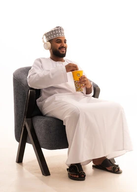 Getting ready to watch movies and bringing delicious snacks, watching a cinematic film, a portrait of an Arab Omani Gulf man wearing a dishdasha and a kumma, with headphones sitting on a chair holding a box of popcorn, gestures of happiness and enjoyment, white background.