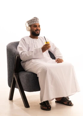 إيماءات السعادة و السرور، الاستمتاع بمشاهدة الأفلام، الحصول على عصير صيفي بارد ومنعش، بورتريه لرجل عربي عماني خليجي يرتدي الدشداشة و الكمة و سماعات الهيدفون ويمسك بيده كأس من المشروب المفضل، خلفية بيضاء