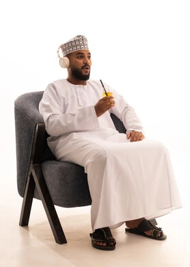 الاستمتاع بمشاهدة الأفلام، إيماءات المفاجأة والاندماج، الحصول على عصير صيفي بارد ومنعش، بورتريه لرجل عربي عماني خليجي يرتدي الدشداشة و الكمة و سماعات الهيدفون ويمسك بيده كأس من المشروب المفضل، خلفية بيضاء
