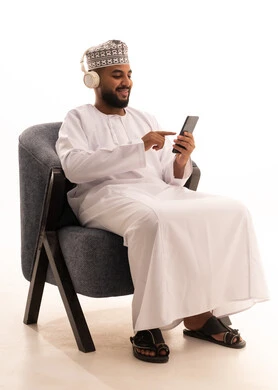Chatting and communicating with family and friends, browsing mobile applications, a portrait of an Arab Omani Gulf man wearing a dishdasha and kumma and headphones sitting on a chair following social media news using a mobile phone, white background.