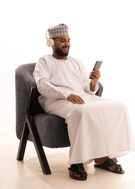 Browsing mobile applications, chatting and talking with friends online, a portrait of an Arab Omani Gulf man wearing a dishdasha and kumma and headphones sitting on a chair following social media news using a mobile phone, white background.