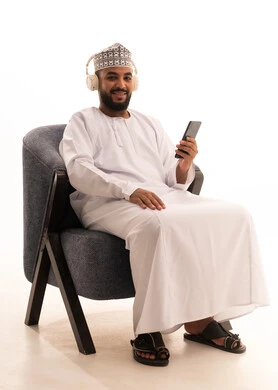 Chatting and talking with friends online, and a portrait of an Arab Omani Gulf man wearing a dishdasha and a kumma, with headphones sitting on a chair following social media news using a mobile phone, browsing mobile applications, white background.