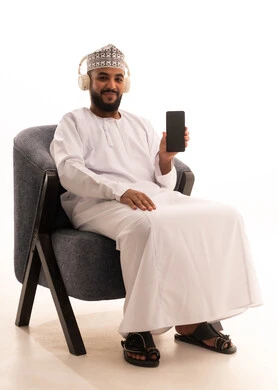A display of a blank black screen on a mobile phone, using advanced modern technology and devices, a portrait of an Arab Omani Gulf man wearing a dishdasha and kumma and headphones sitting on a chair looking at the camera with gestures of joy, white background.