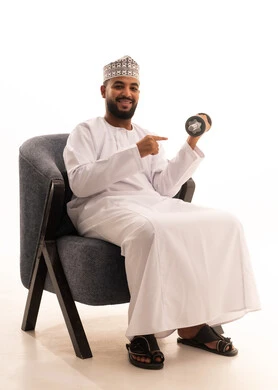 نشاط بدني للمحافظة على اللياقة الجسدية، رفع الأوزان الحديدية الثقيلة، بناء عضلات الجسم وتقويتها، بورتريه لرجل عربي عماني خليجي يرتدي الدشداشة و الكمة يجلس على كرسي يحمل بيده الأثقال و يشير اليها ، الاهتمام باللياقة والصحة الجسدية، خلفية بيضاء