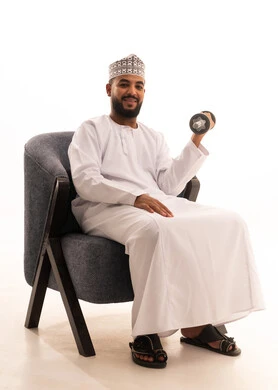 رفع الأوزان الحديدية الثقيلة، نشاط بدني للمحافظة على اللياقة الجسدية، بناء عضلات الجسم وتقويتها، بورتريه لرجل عربي عماني خليجي يرتدي الدشداشة و الكمة يجلس على كرسي يحمل بيده الأثقال، الاهتمام باللياقة والصحة الجسدية، خلفية بيضاء