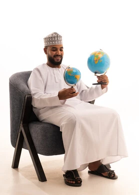 تعابير السرور و الفرح بالانتماء للدول العربيه, مفهوم حب البحث واستكشاف القارات في الجغرافيا, بورتريه لرجل عربي عماني خليجي يرتدي الدشداشة و الكمة يجلس على كرسي يحمل بيده مجسم الكرة الارضية، خلفيه بيضاء