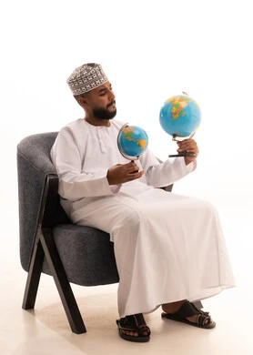 ايماءات التركيز ومفهوم البحث والاستكشاف حول العالم, بورتريه لرجل عربي عماني خليجي يرتدي الدشداشة و الكمة يجلس على كرسي يحمل بيده مجسم الكرة الارضية, خلفية بيضاء