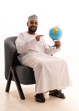 The concept of the love for research and exploring continents in geography, a portrait of an Arab Omani Gulf man wearing a dishdasha and a kumma sitting on a chair holding a globe in his hand, expressions of joy and happiness for belonging to the Arab countries, white background.
