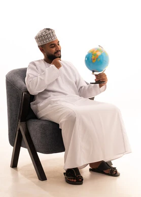 Gestures of finding a specific place on the map and looking at one of the continents, the concept of research and exploration around the world, and a portrait of an Arab Omani Gulf man wearing a dishdasha and a kumma sitting on a chair holding a globe in his hand, with a white background.