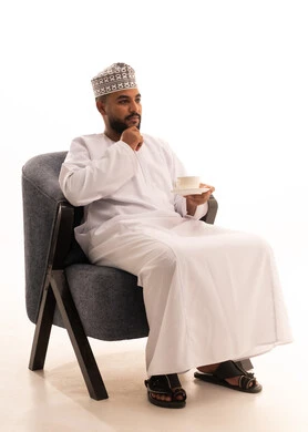 Thinking about something, enjoying hot drinks, spending free time sipping the favorite beverage, a portrait of an Arab Gulf Omani man wearing a dishdasha and a turban sitting on a chair drinking coffee with gestures of happiness and joy, white background.