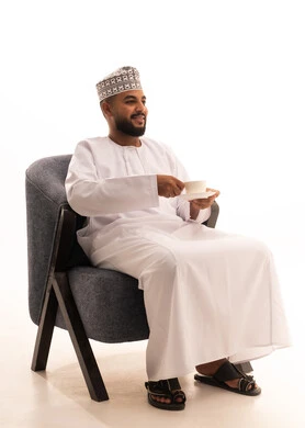 Spending free time sipping a favorite drink, enjoying hot beverages, having warm winter herbs, a portrait of an Arab Gulf Omani man wearing a dishdasha and a turban sitting on a chair drinking coffee with gestures of happiness and joy, white background.