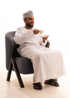Enjoying hot drinks, having warm winter herbs, spending leisure time sipping a favorite beverage, a portrait of an Arab Gulf Omani man wearing a dishdasha and turban sitting on a chair drinking coffee with gestures of happiness and joy, white background.