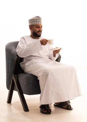 تناول الأعشاب الشتوية الدافئة، الاستمتاع بالمشروبات الساخنة، قضاء وقت الفراغ باحتساء المشروب المفضل, بورتريه لرجل عربي خليجي عماني يرتدي الدشداشة والعمامة يجلس على كرسي يقوم بشرب القهوة بإيماءات السعادة والسرور، خلفية بيضاء