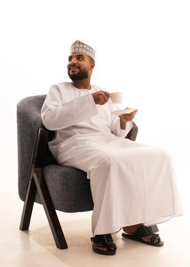 النظر الى شيء ما، الاستمتاع بالمشروبات الساخنة، قضاء وقت الفراغ باحتساء المشروب المفضل, بورتريه لرجل عربي خليجي عماني يرتدي الدشداشة والعمامة يجلس على كرسي يقوم بشرب القهوة بإيماءات السعادة والسرور، خلفية بيضاء