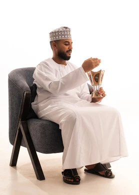 التبخير واستخدام العطور والمطيبات، اتباع العادات والتقاليد العربية الأصيلة، التبخر في الأعياد والمناسبات السعيدة،بورتريه لرجل عربي خليجي عماني يرتدي الكمه والدشداشة يجلس على كرسي ويمسك بيده المبخرة، الطول الكامل للجسم، خلفية بيضاء