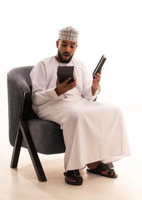 مفهوم المفاجأة والإهداء، إيماءات السعادة والدهشة بالحصول على هدية، فن انتقاء الهدايا بعناية، بورتريه لرجل عربي خليجي عماني يرتدي الكمه والدشداشة يجلس على كرسي ويقوم بفتح العلبة بتعابير الفرح والسرور، خلفية بيضاء