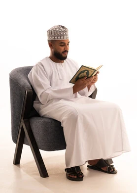 The feeling of humility and tranquility, reciting and reading the Holy Quran and contemplating its meanings, a portrait of an Arab Gulf Omani man wearing a kumma and dishdasha sitting on a chair holding the Holy Quran in his hand, frequently mentioning Allah Almighty, white background.