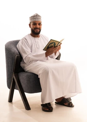 Recitation and reading of the Holy Quran and contemplation of its meanings, a portrait of an Arab Gulf Omani man wearing a kumma and dishdasha sitting on a chair holding the Holy Quran in his hand, frequent remembrance of Allah Almighty, feeling of humility and tranquility, white background.