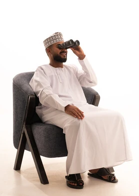 إيماءات الاستمتاع والسرور، حب الاستكشاف والمغامرة، استعمال المنظار للكشف عن الاشياء البعيدة وتقريبها, بورتريه لشاب عربي خليجي عماني يرتدي الكمه والدشداشة يجلس على كرسي مريح يحمل بيده المنظار ثنائي العينين وينظر الى شيء ما، خلفية بيضاء