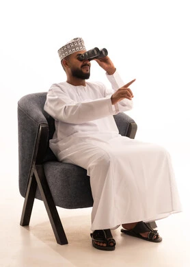 حب الاستكشاف والمغامرة، استعمال المنظار للكشف عن الاشياء البعيدة وتقريبها, بورتريه لشاب عربي خليجي عماني يرتدي الكمه والدشداشة يجلس على كرسي مريح يحمل بيده المنظار ثنائي العينين وينظر الى شيء ما، إيماءات الاستمتاع والسرور، خلفية بيضاء