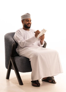 تدوين وتسجيل الملاحظات والمعلومات على الورق، تنظيم وإدارة الأعمال , بورتريه لرجل عربي عماني يرتدي الدشداشة والكمه يجلس على كرسي يقوم بكتابة المهام والأهداف اليومية، خلفية بيضاء