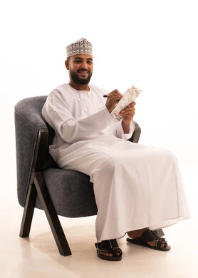 تنظيم وإدارة الأعمال ، تدوين وتسجيل الملاحظات والمعلومات على الورق, بورتريه لرجل عربي عماني يرتدي الدشداشة والكمه يجلس على كرسي يقوم بكتابة المهام والأهداف اليومية، خلفية بيضاء