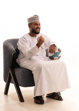 Developing sports hobbies, raising a fist in gestures of victory, watching and following a football match, gestures of passion and encouragement for the team, a portrait of an Omani Arab man wearing a dishdasha and kumma sitting on a chair holding a ball, enthusiasm and interaction with the national team, white background.