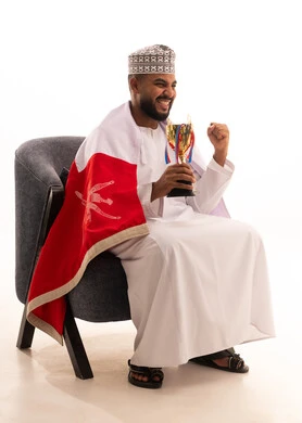 إيماءات الشغف والتشجيع للفريق، رفع قبضة اليد بايماءات الحماس والتفاعل مع المنتخب، مشاهدة ومتابعة مباراة كرة القدم، بورتريه لرجل عربي عماني يرتدي الدشداشة والكمه يجلس على كرسي يحمل كأس المنتخب، أنديه رياضية مختلفة، خلفية بيضاء