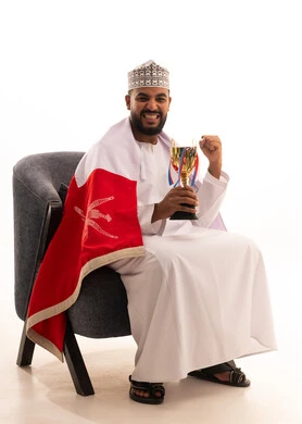 رفع قبضة اليد بايماءات الحماس والتفاعل مع المنتخب، إيماءات الشغف والتشجيع للفريق، مشاهدة ومتابعة مباراة كرة القدم، بورتريه لرجل عربي عماني يرتدي الدشداشة والكمه يجلس على كرسي يحمل كأس المنتخب، أنديه رياضية مختلفة، خلفية بيضاء