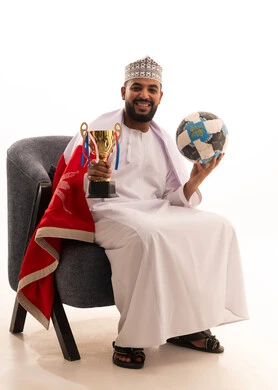 تشجيع الأنديه والمنتخبات، ايماءات السعادة و السرور، الشغف في مباريات كرة القدم، بورتريه لرجل عربي عماني يرتدي الدشداشة والكمه يجلس على كرسي يحمل بيدية الكرة وكأس الفوز، خلفية بيضاء