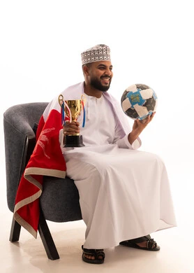 الشغف في مباريات كرة القدم، بورتريه لرجل عربي عماني يرتدي الدشداشة والكمه يجلس على كرسي يحمل بيدية الكرة وكأس الفوز، تشجيع الأنديه والمنتخبات، خلفية بيضاء