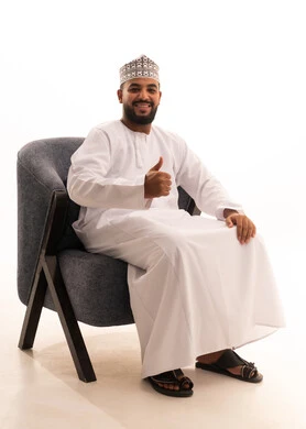 علامة الجودة والامتياز, بورتريه لرجل عربي عماني يرتدي الدشداشة والكمه يجلس على كرسي يرفع ابهامه بإيماءات الموافقة والقبول, الاعجاب والرضا لتحقيق أمر معين, خلفية بيضاء