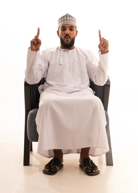 Gestures of astonishment and amazement, pointing and clarifying something, a portrait of an Omani Arab man wearing a dishdasha and kumma sitting on a chair pointing with his index fingers upwards with expressions of this place here, specifying a certain location, white background.