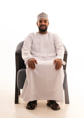 مفهوم القوة والرجولة والشياكة، ايماءات الثقة بالنفس، الاهتمام بالأناقة والمظهر الخارجي، بورتريه لرجل عربي عماني يرتدي الدشداشة والكمه يجلس على كرسي ينظر الى الكاميرا بتعابير الفرح والسرور، خلفية بيضاء