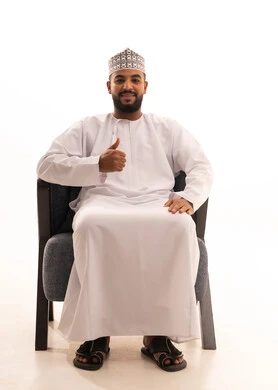 الإمتياز والتميز والنجاح، بورتريه لرجل عربي عماني يرتدي الدشداشة والكمه يجلس على كرسي ويرفع أصابع الإبهام لأعلى، إيماءات السعادة والإعجاب والموافقة، حركة أصبع تدل على القبول والرضا، خلفية بيضاء