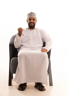 مفهوم الانجاز وتحقيق الاهداف، الشعور بالسعادة والقوة، بورتريه لرجل عربي عماني يرتدي الدشداشة والكمه يجلس على كرسي ينظر الى الكاميرا يرفع قبضة يده للاعلى بإيماءات الفرح والسرور, الانتصار والفوز والنجاح، خلفية بيضاء